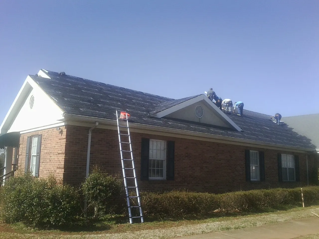 Crew tearing shingles off a brick ranch roof with a ladder set — Metal Roof Repair work by our crew in Alden