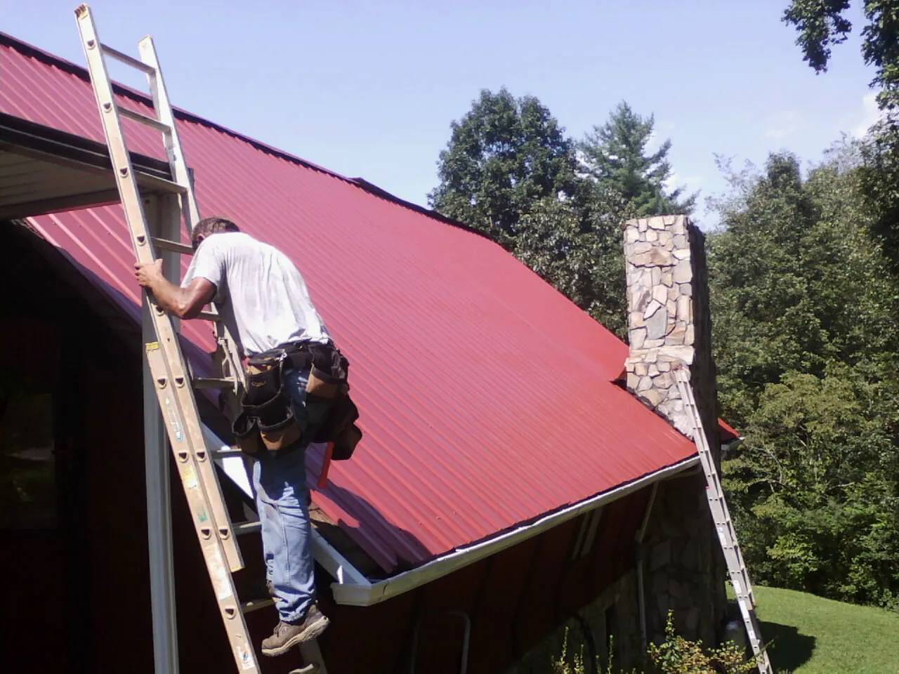 Roofer on a ladder beside a red metal roof and stone chimney — Soffit & Fascia Repair by Roofing Brothers in Alden